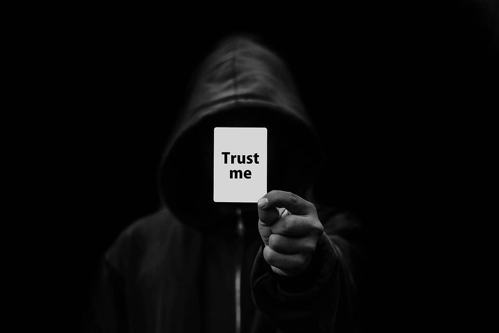 Hooded man in darkness holding a 'trust me' sign